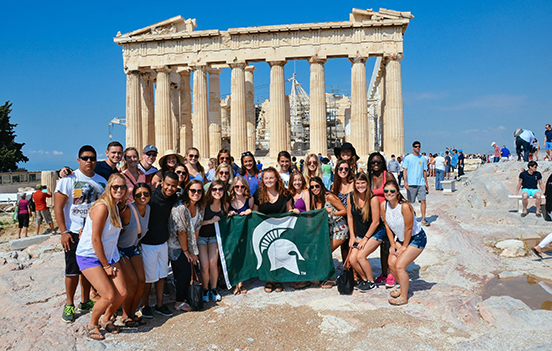 a large group of study abroad students in front of the Acropolis, holding an MSU flag