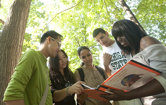 Five students outside on a sunny day looking down at a book